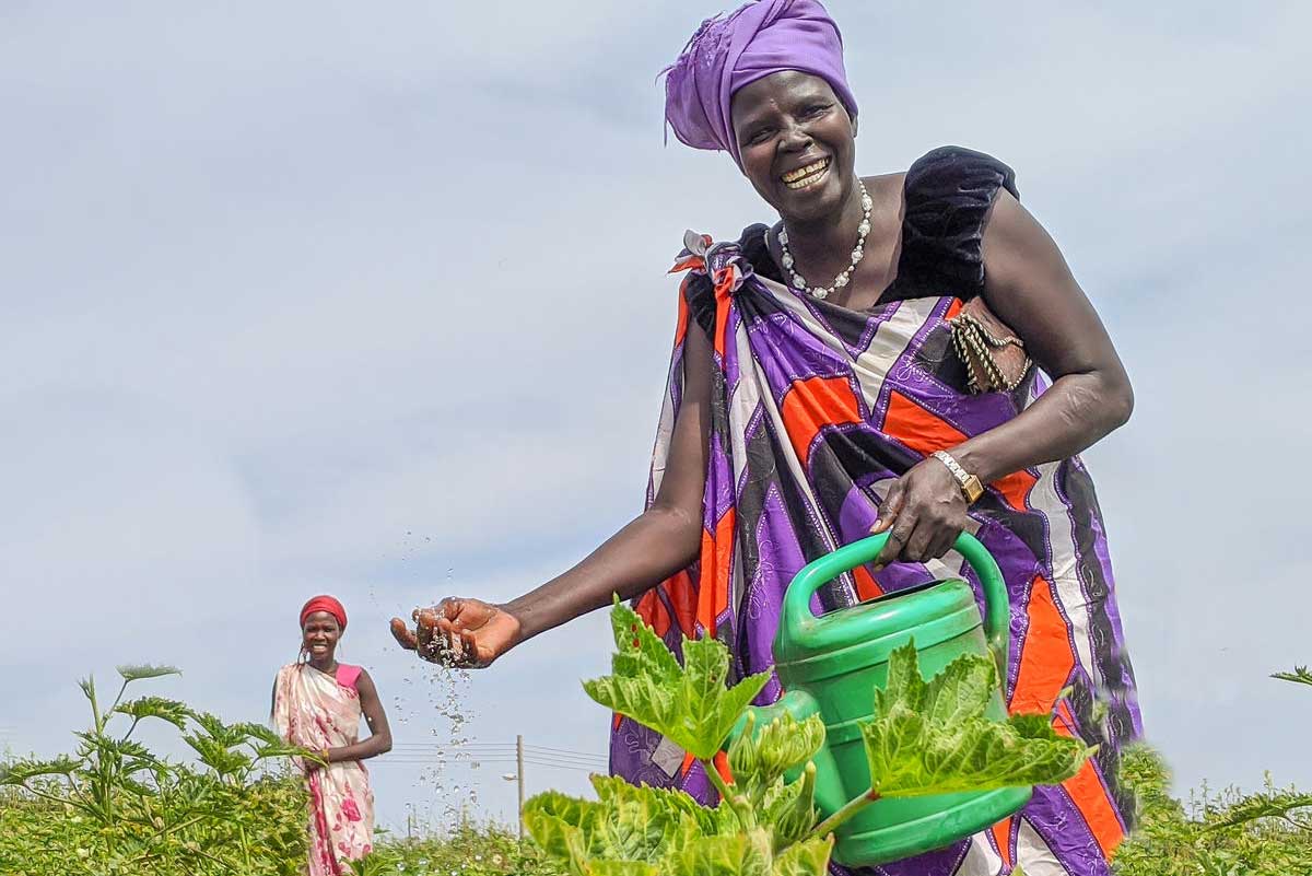 Au Soudan du Sud, une femme au grand sourire arrose un jardin florissant. Une autre femme sourit en arrière-plan.