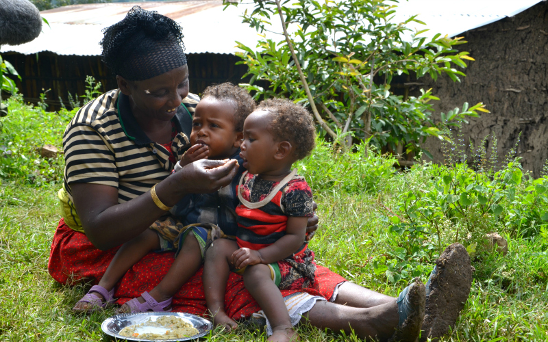 A woman holds two little children on her lap and feeds them