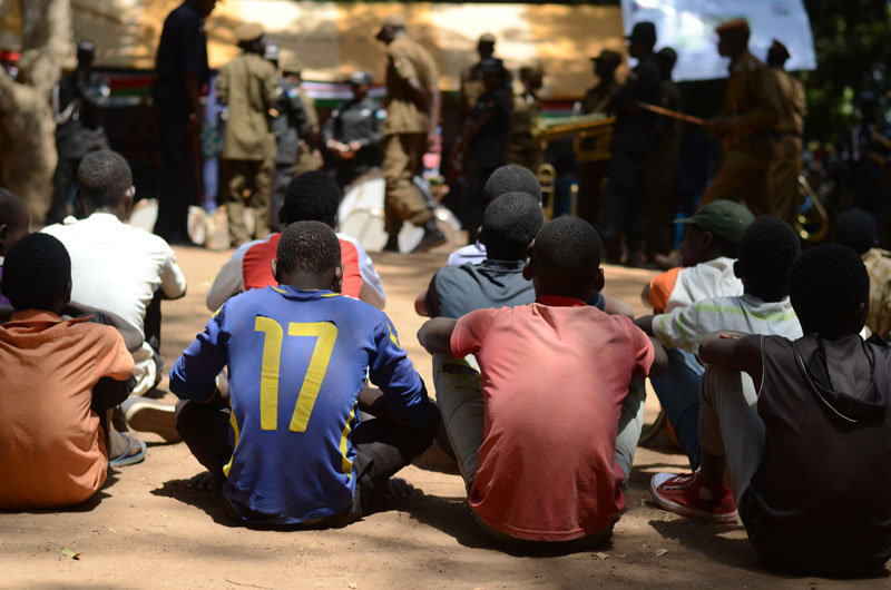Former child soldier teenage boys appear sitting on a dirt ground with their backs to the camera, waiting to receive assistance.