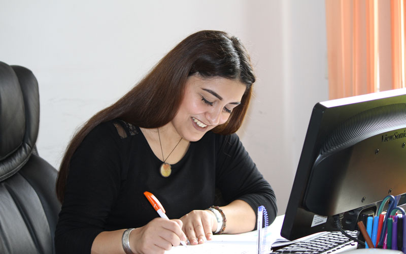 Une femme en tenue professionnelle est assise à son bureau et écrit en souriant.