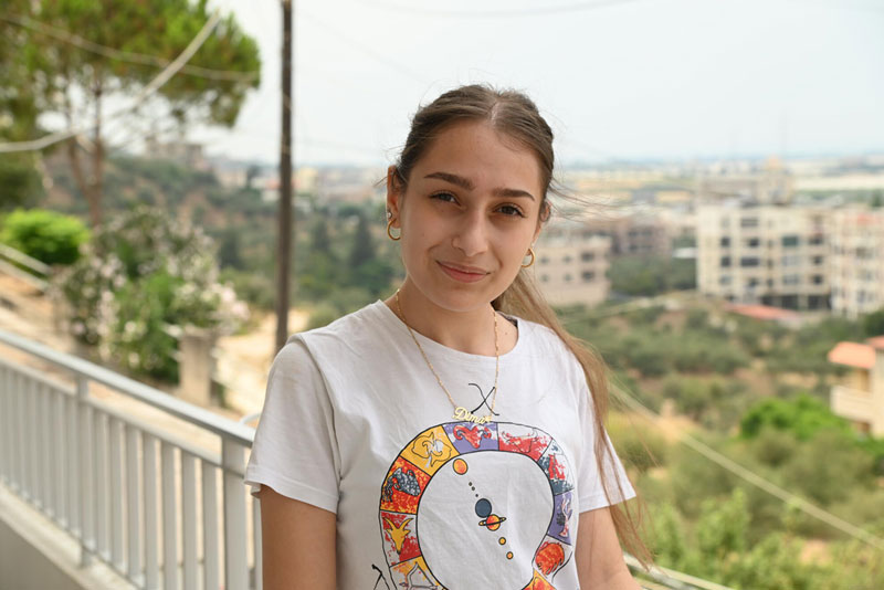 Une adolescente debout devant une balustrade de balcon porte un t-shirt blanc et sourit pour la photo.
