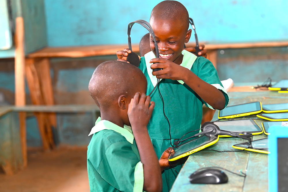 Deux enfants souriants en uniforme vert mettent des écouteurs et interagissent avec des tablettes posées sur une table, dans une salle de classe.