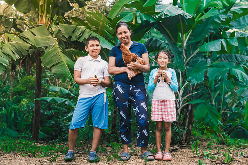 Une femme, une poule dans les bras, se tient entre ses deux enfants souriants, chacun tenant un poussin, à l’extérieur. Ils sont entourés de feuilles de bananier et de verdure.