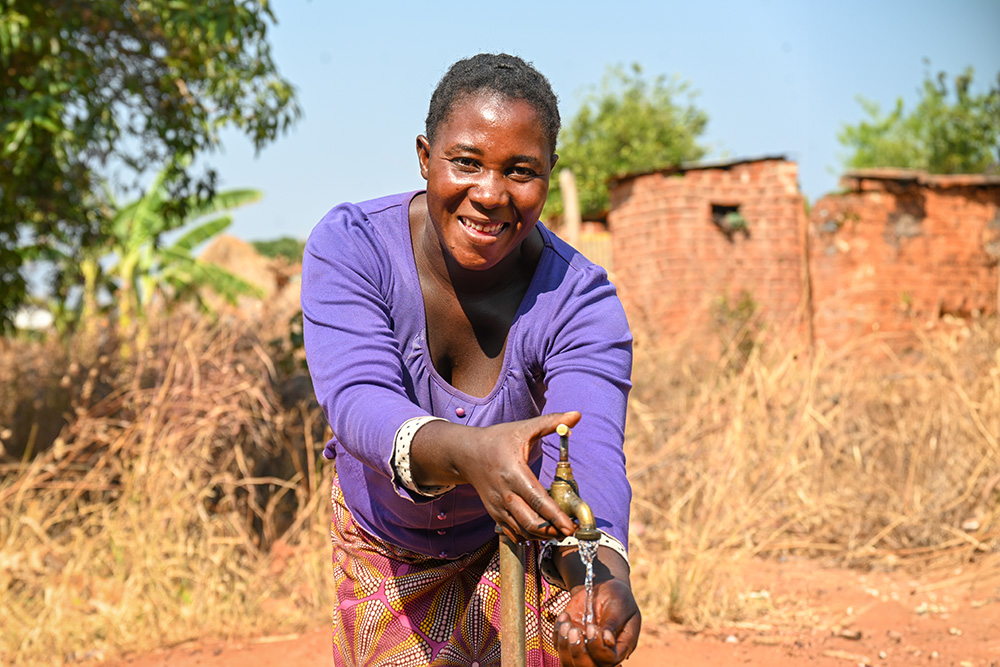 Une femme vêtue d’un haut violet sourit alors qu’elle recueille de l’eau propre à un robinet.