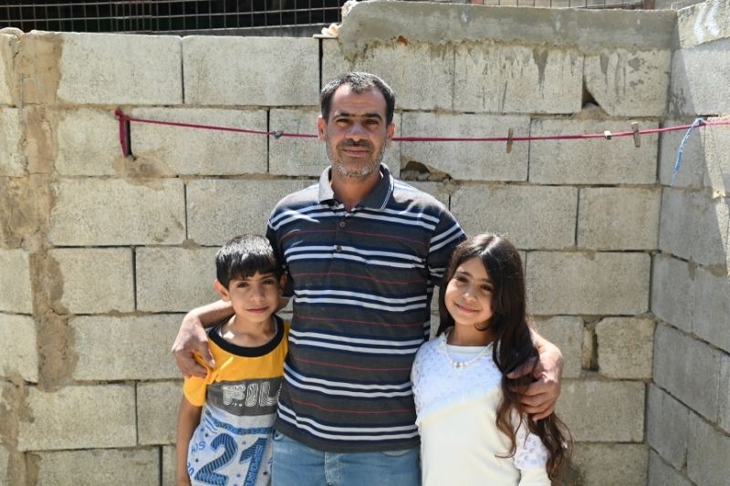 A Lebanese man in a striped polo shirt stands with his arms around a young boy and a young girl.