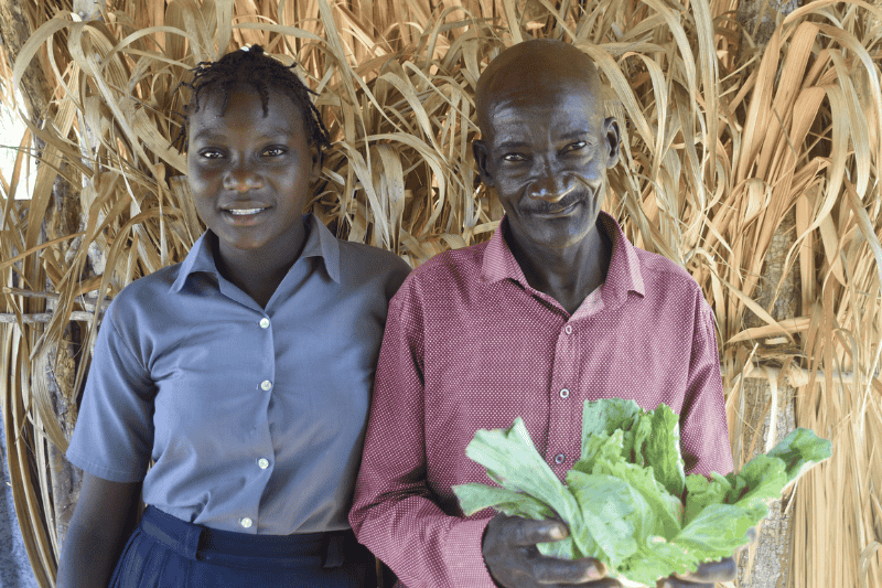 In La Belle-Mere, Haiti, a young girl in a button-down shirt stands with her arm around an older man, who holds green lettuce in his hands.