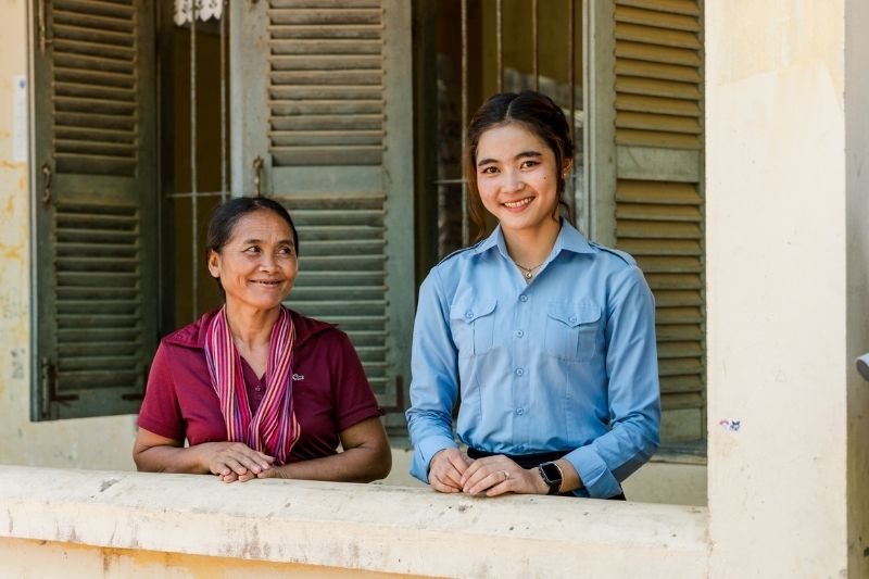  An older Cambodian woman in burgundy beams up at her smiling daughter, who wears a blue button-down shirt.