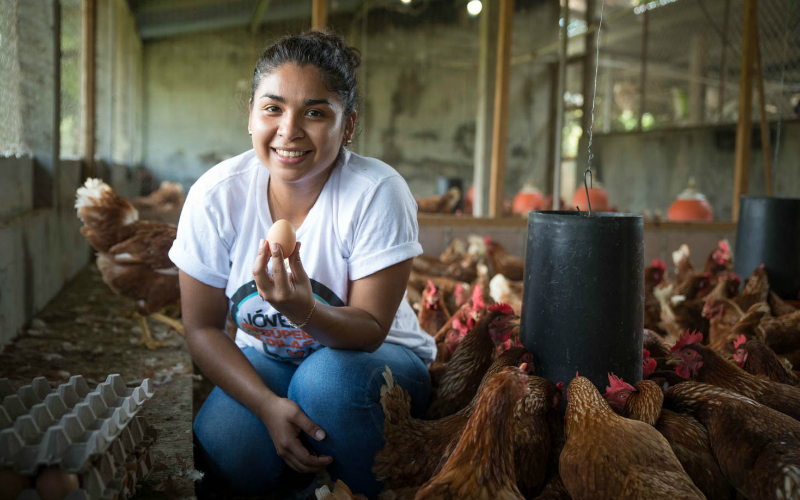 Inside a hen house, a young woman shows off an egg that she has collected.