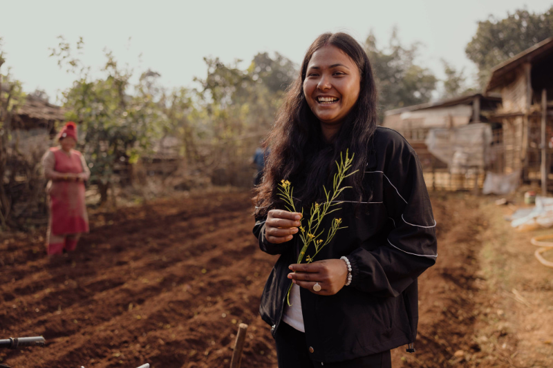 Une jeune femme souriante tient des brins de plante dans un champ fraîchement labouré.